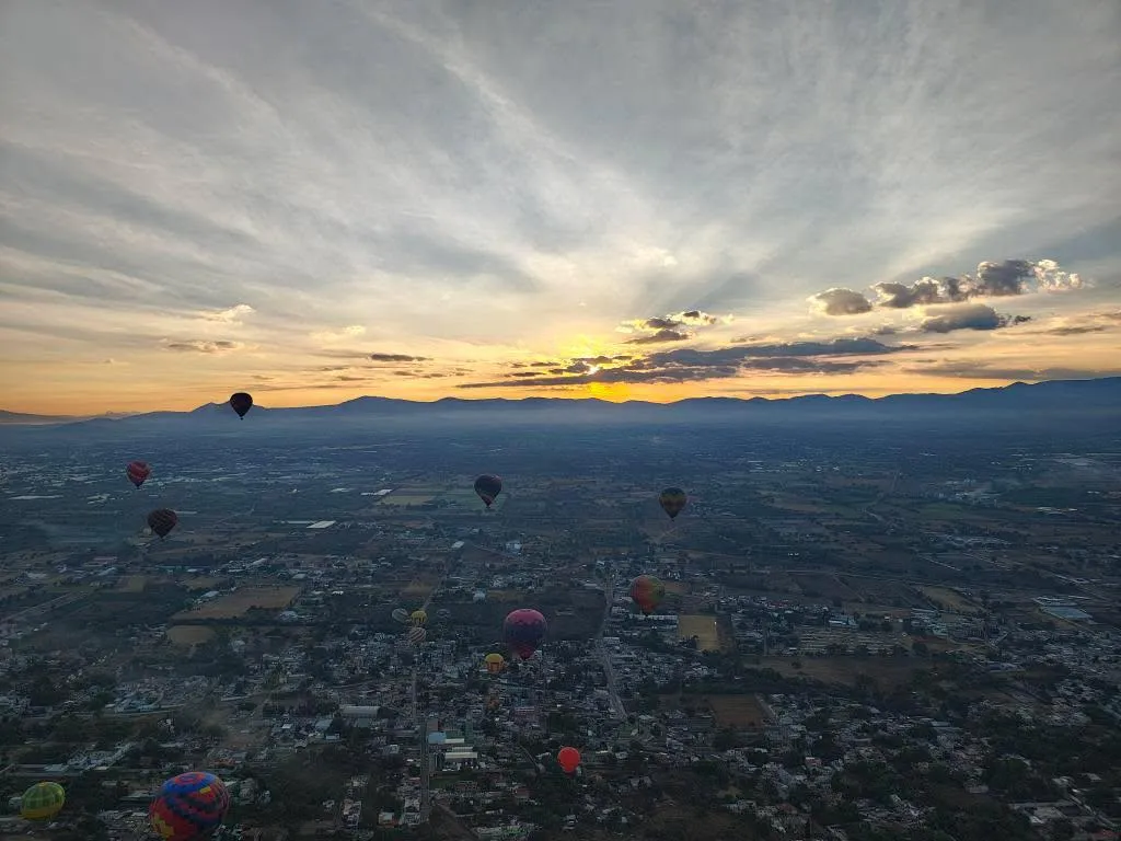 Amanecer entre nubes sobre la sierra con una docena de globos dispersos volando encima del pueblo