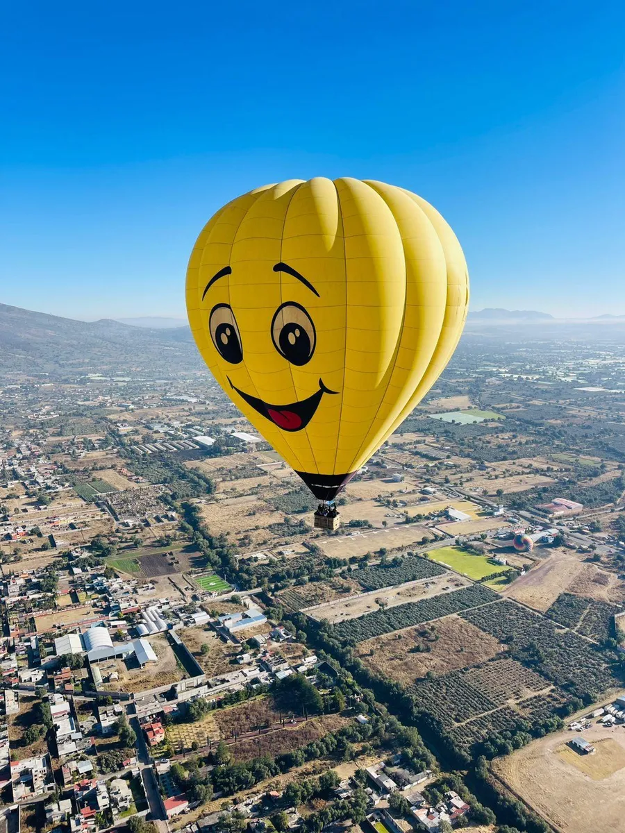 Globo aerostatico amarillo con una carita feliz pintada volando sobre casas y sembradios del valle