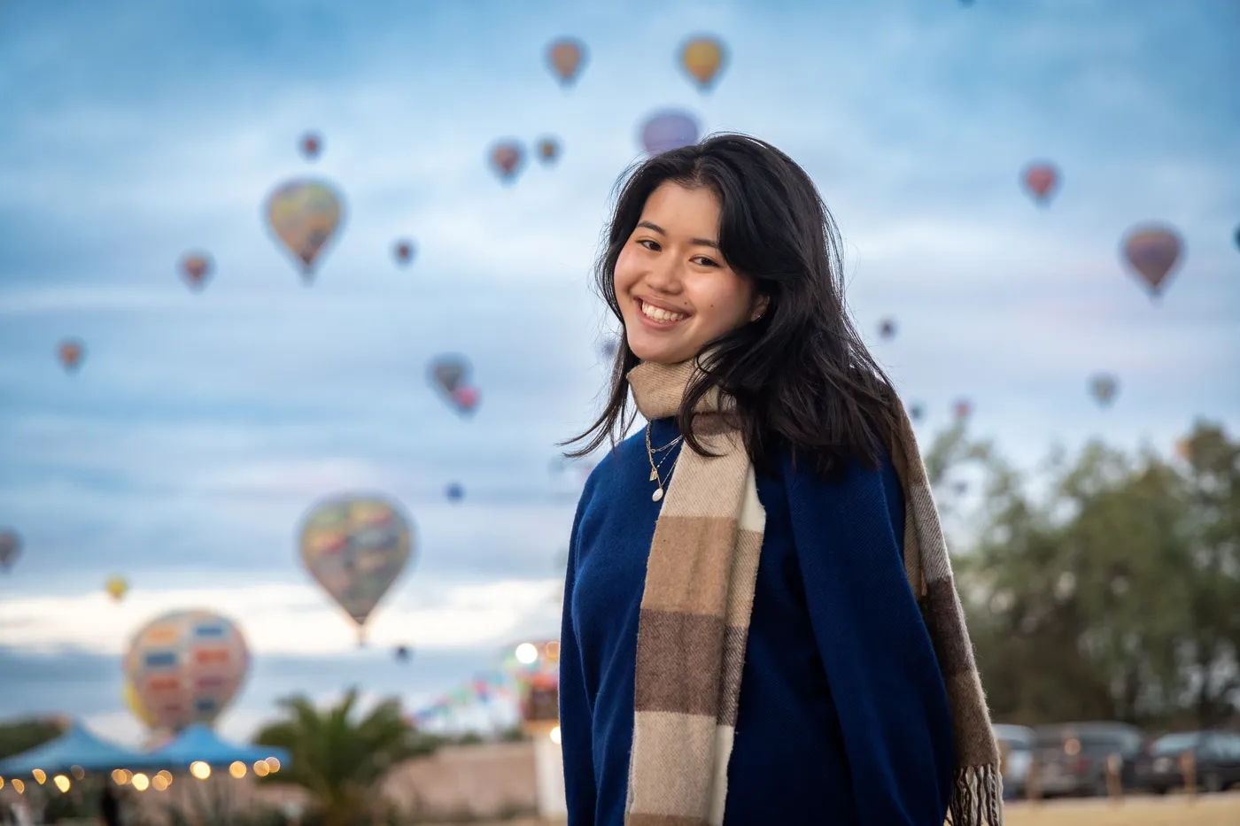 Joven sonriente con bufanda a cuadros frente a decenas de globos aerostáticos al amanecer