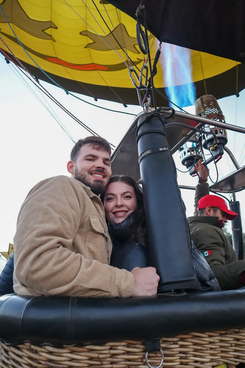 Pareja abrazada en la canastilla mientras el piloto enciende el quemador bajo un globo amarillo