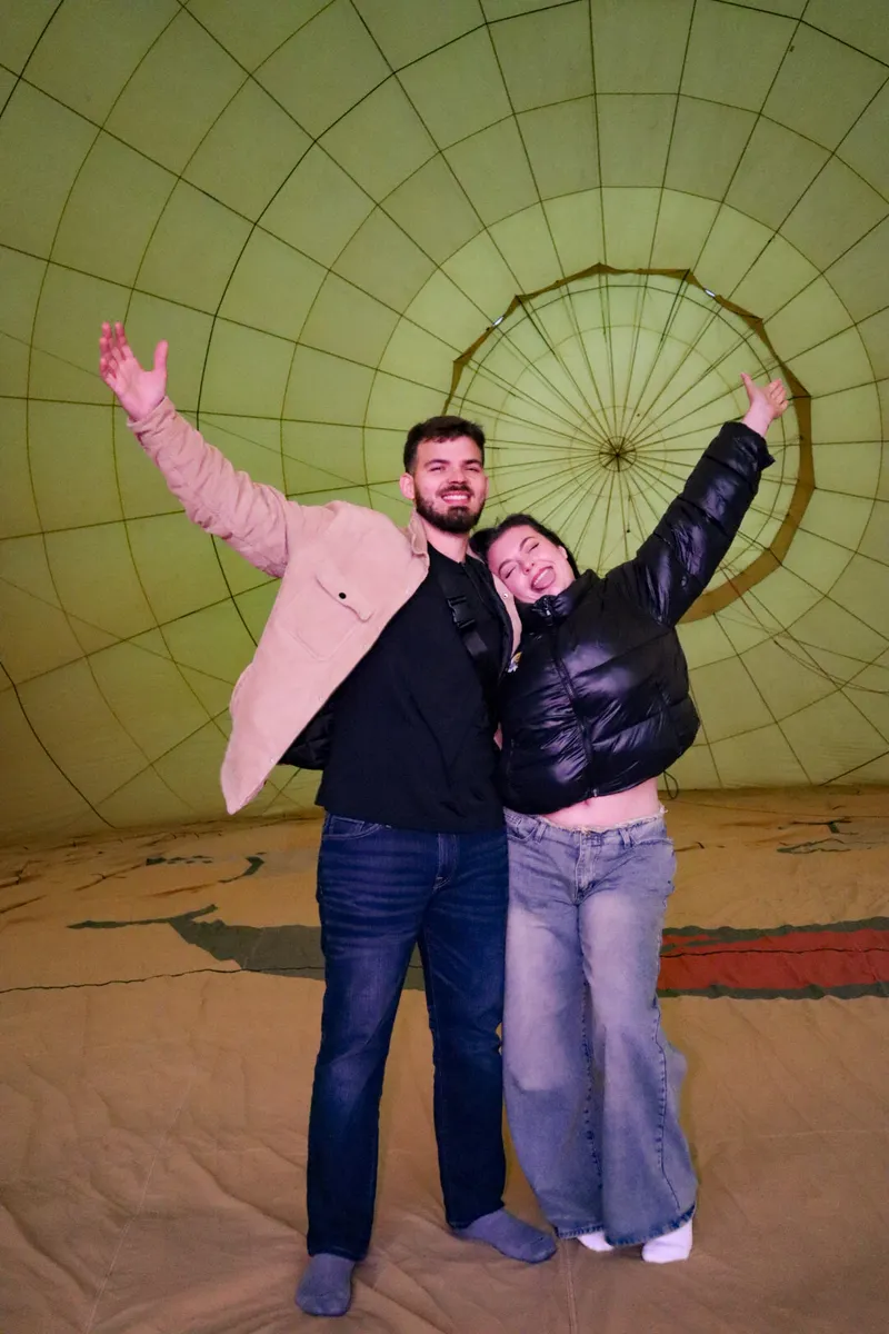 Pareja con los brazos en alto celebra de pie dentro de la lona verde del globo ya inflado antes del vuelo
