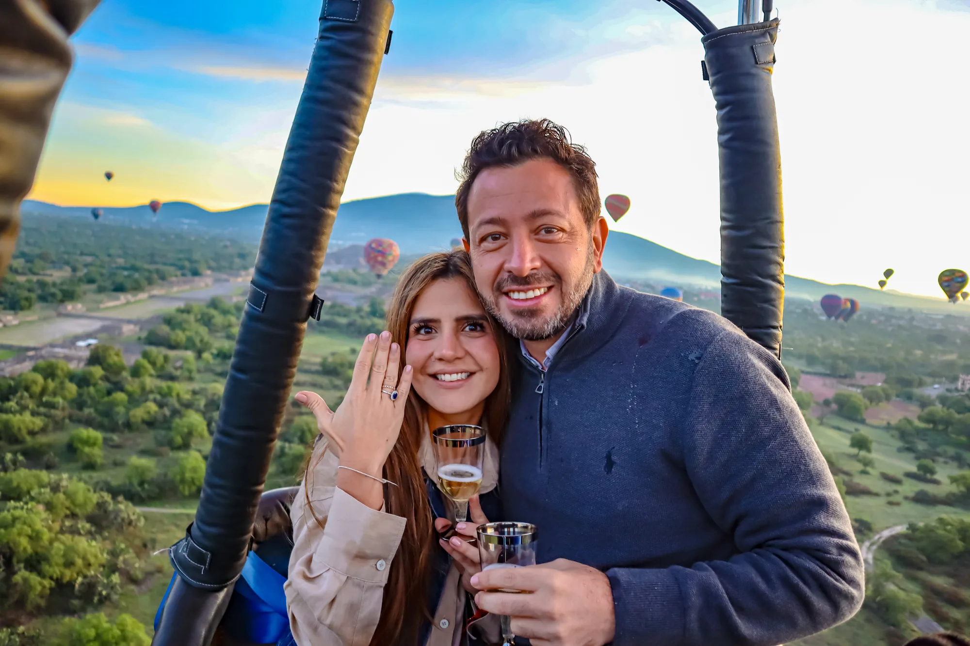 Pareja recién comprometida brindando tras la entrega del anillo, con globos aerostáticos al amanecer sobre Teotihuacán